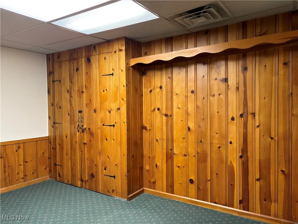 Empty room featuring a wainscoted wall, carpet flooring, wooden walls, visible vents, and a drop ceiling