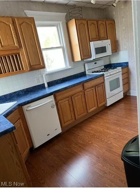 Kitchen featuring backsplash, brown cabinets, white appliances, dark countertops, and dark wood-type flooring