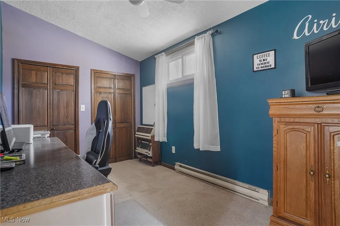 Home office with vaulted ceiling, a textured ceiling, ceiling fan, a baseboard radiator, and light carpet