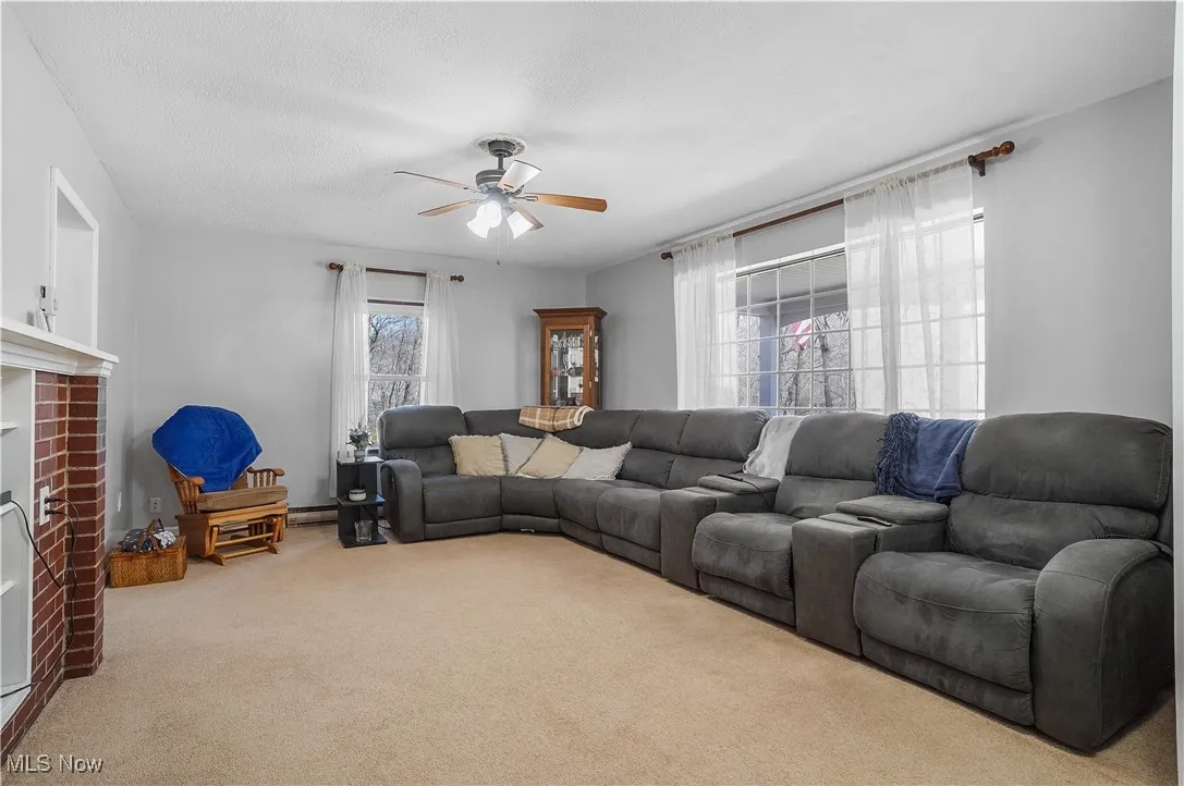 Carpeted living area featuring baseboard heating, a textured ceiling, a ceiling fan, and a brick fireplace