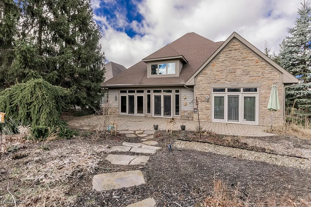 Back of property with stone siding, a shingled roof, and a patio