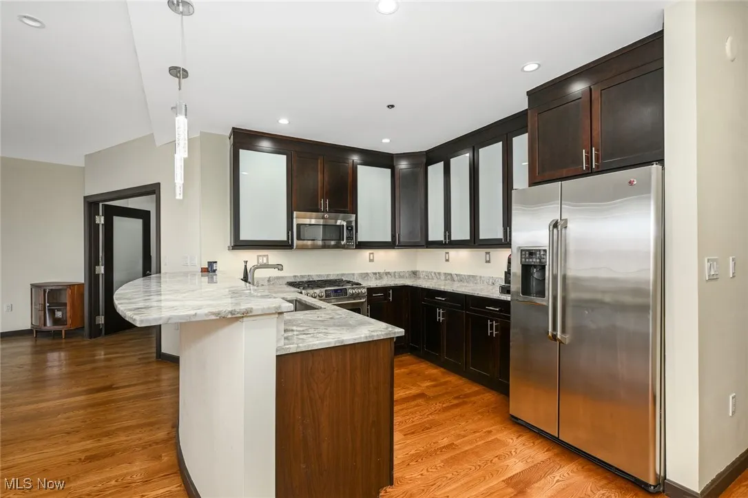 Kitchen with a peninsula, light wood finished floors, light stone countertops, appliances with stainless steel finishes, and hanging light fixtures