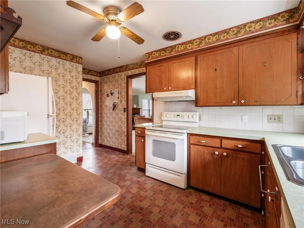 Kitchen with white appliances, ceiling fan, wallpapered walls, under cabinet range hood, and a sink