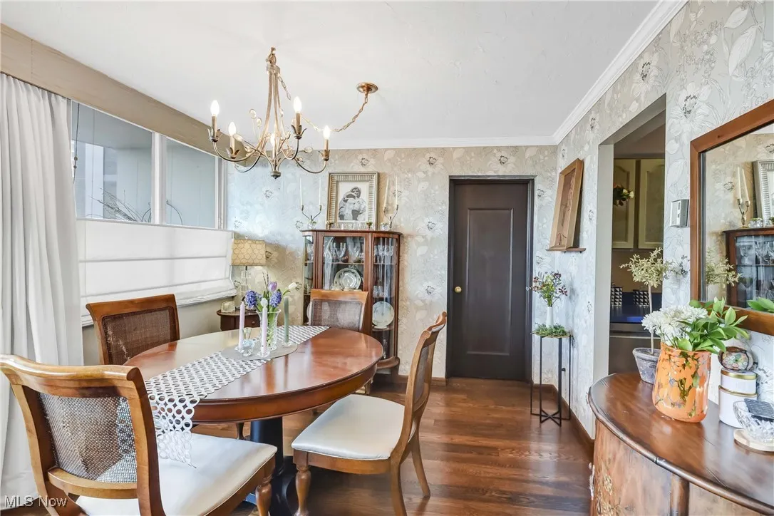 Dining space with dark wood-type flooring, wallpapered walls, an inviting chandelier, and crown molding