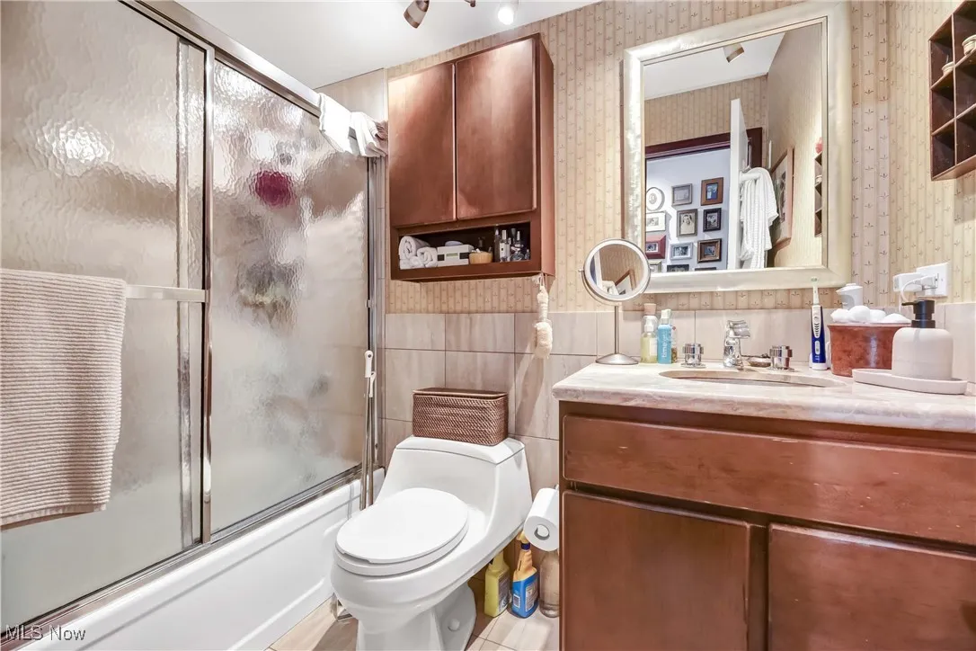 Full bath with combined bath / shower with glass door, toilet, vanity, wallpapered walls, and tile walls