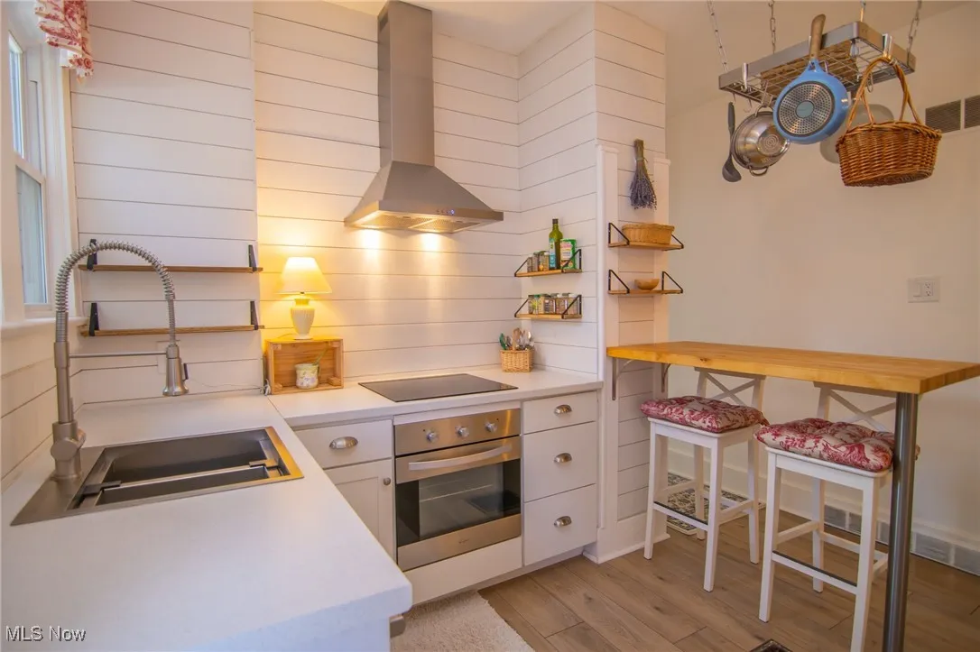 Kitchen featuring light countertops, stainless steel oven, a sink, black electric stovetop, and wall chimney range hood