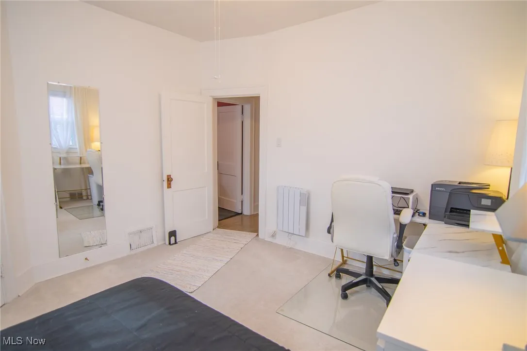 Carpeted home office featuring baseboards, visible vents, and radiator heating unit