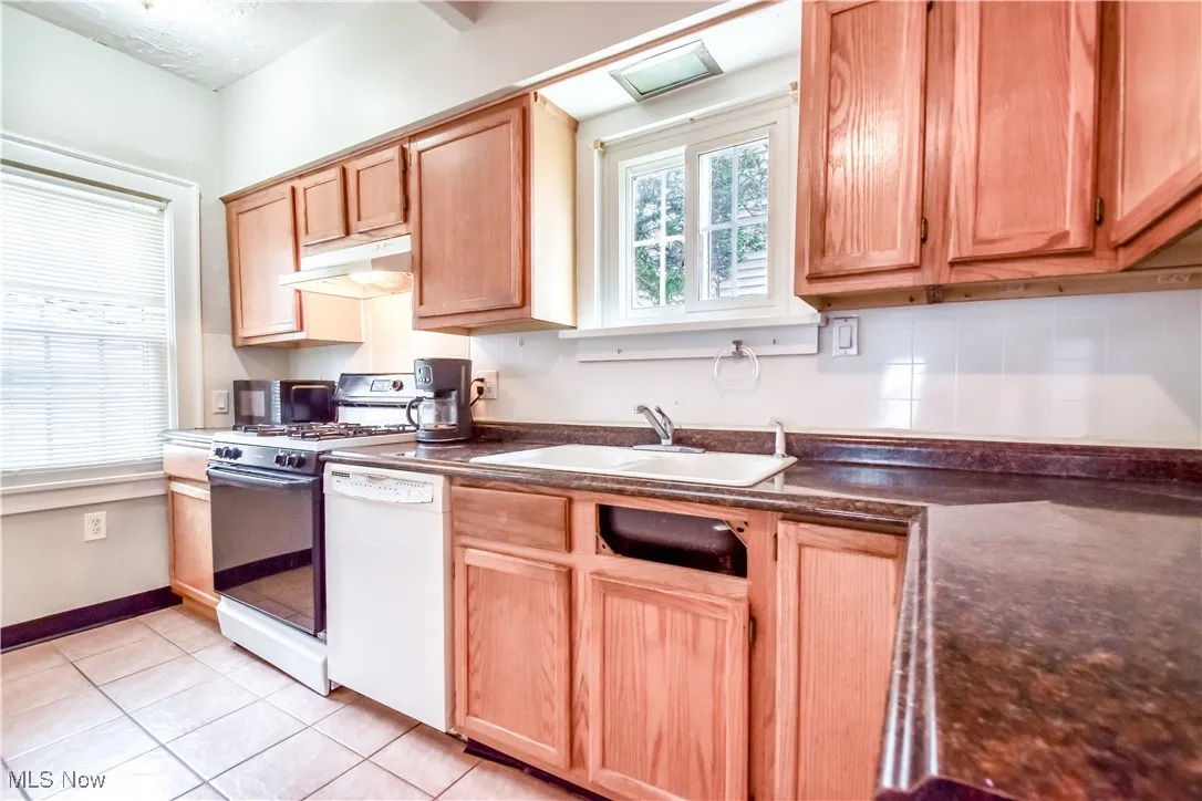 Kitchen featuring white appliances, light tile patterned floors, under cabinet range hood, a sink, and dark countertops