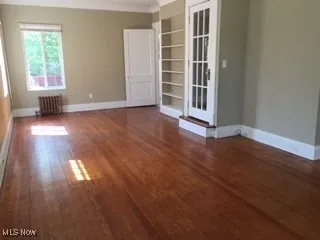 Empty room with crown molding, baseboards, built in shelves, radiator heating unit, and wood finished floors