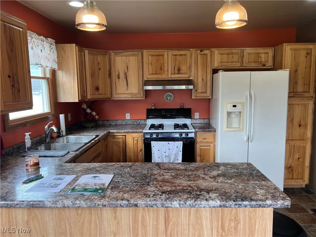 Kitchen featuring white fridge with ice dispenser, gas stove, under cabinet range hood, hanging light fixtures, and dark stone counters