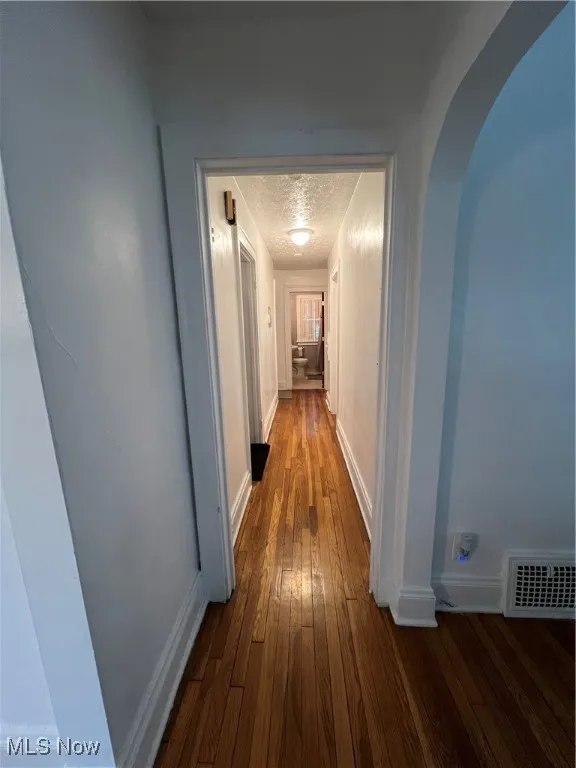 Corridor featuring baseboards, a textured ceiling, dark wood-type flooring, visible vents, and arched walkways