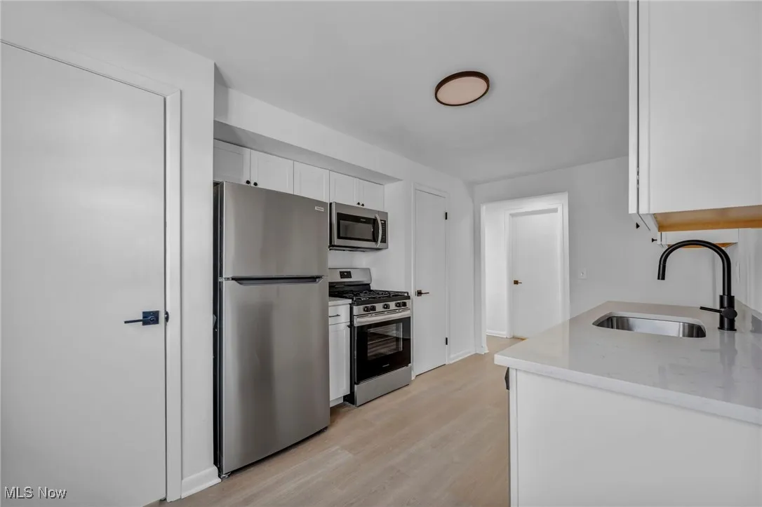 Kitchen with a sink, appliances with stainless steel finishes, white cabinetry, light wood-style flooring, and light stone counters