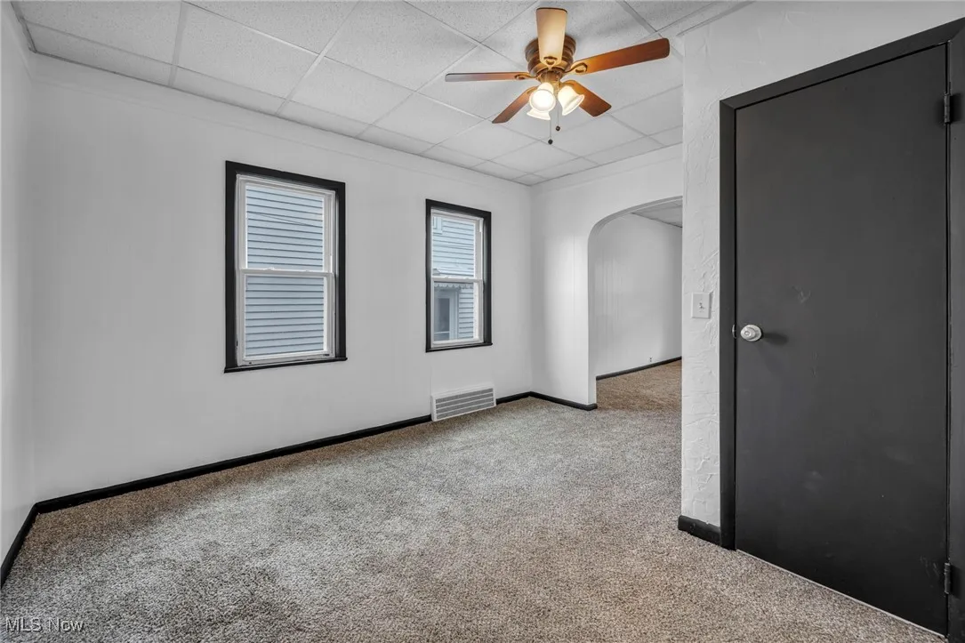 Carpeted empty room featuring a paneled ceiling, arched walkways, a ceiling fan, visible vents, and baseboards