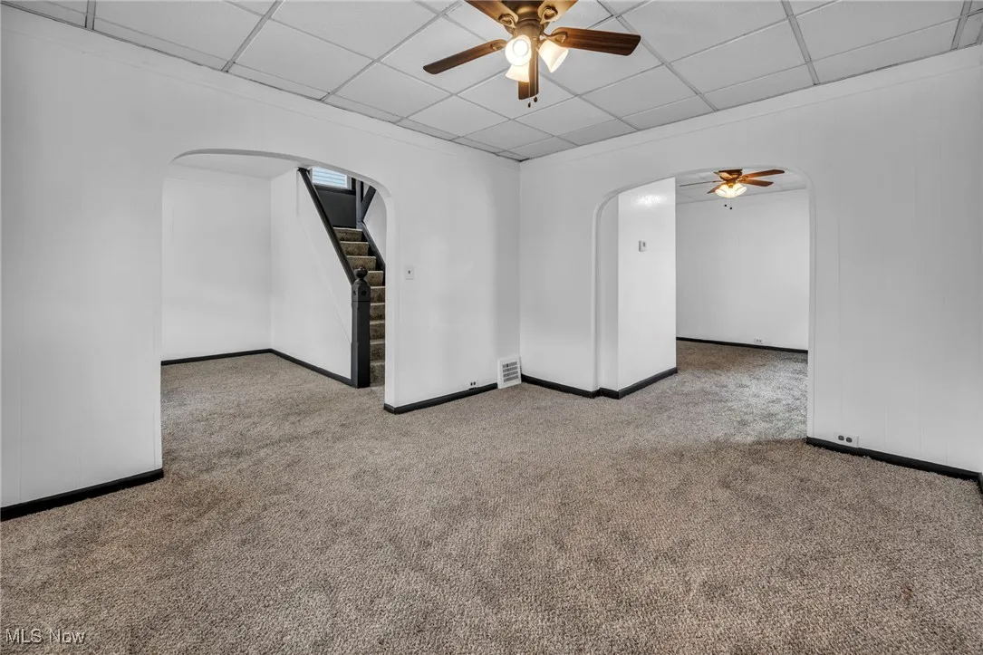Carpeted empty room featuring arched walkways, a paneled ceiling, a ceiling fan, and stairway
