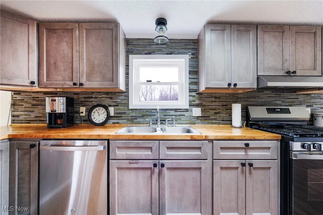 Kitchen with butcher block countertops, backsplash, under cabinet range hood, appliances with stainless steel finishes, and a sink