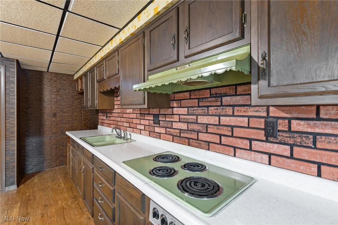 Kitchen featuring a paneled ceiling, dark brown cabinetry, electric stovetop, under cabinet range hood, and a sink