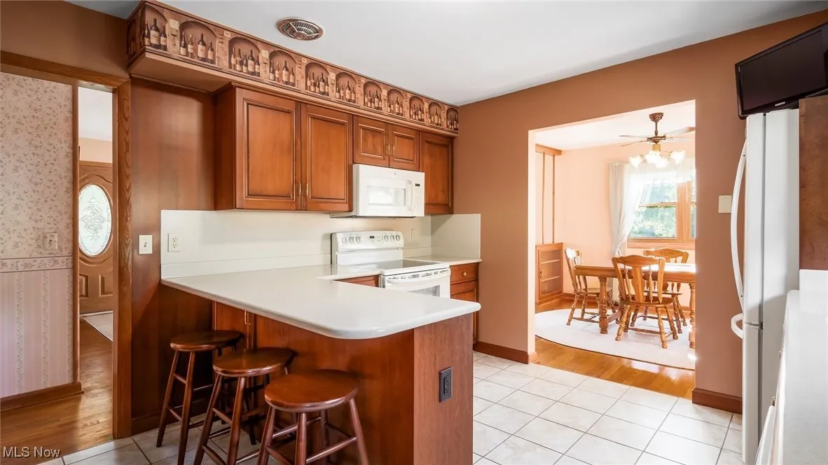 Kitchen with a kitchen breakfast bar, white appliances, a peninsula, brown cabinetry, and wallpapered walls