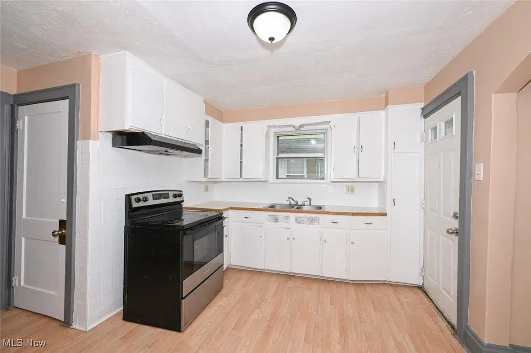 Kitchen with light wood-type flooring, stainless steel electric range, white cabinets, under cabinet range hood, and a sink