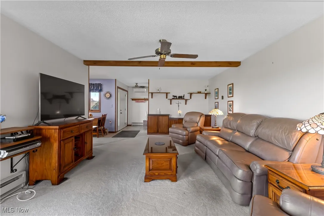 Living room with baseboard heating, light carpet, a textured ceiling, a ceiling fan, and beamed ceiling