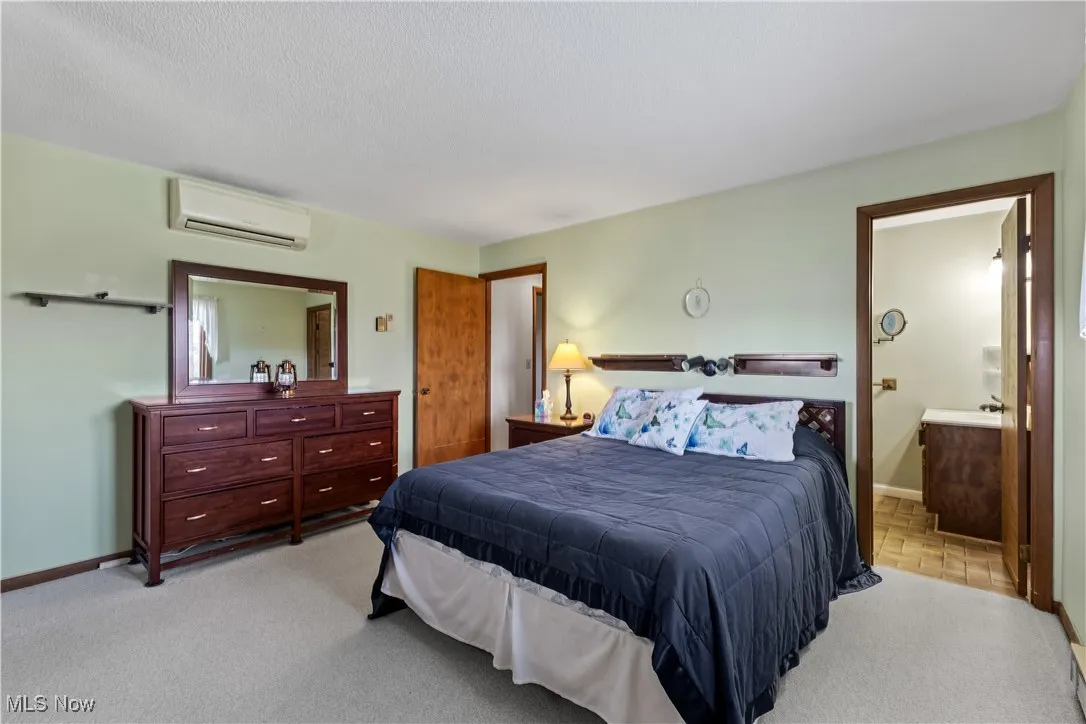 Bedroom featuring ensuite bathroom, baseboards, a wall mounted AC, a textured ceiling, and light colored carpet
