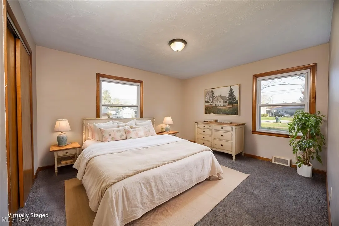 Virtually staged primary bedroom featuring dark carpet, visible vents, baseboards, and multiple windows
