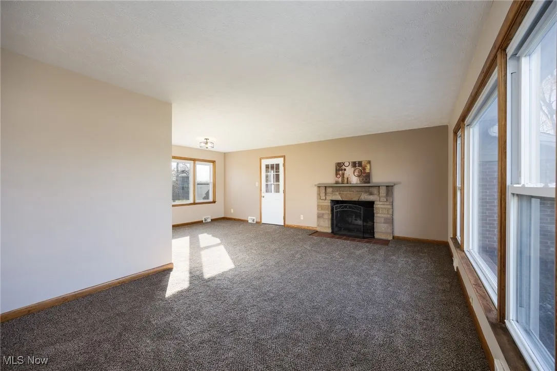 Unfurnished living room with dark carpet, a stone fireplace, and baseboards