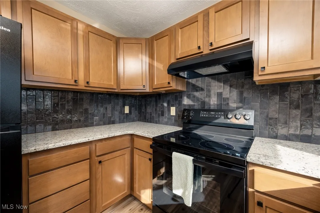 Kitchen with light stone countertops, decorative backsplash, under cabinet range hood, and black appliances