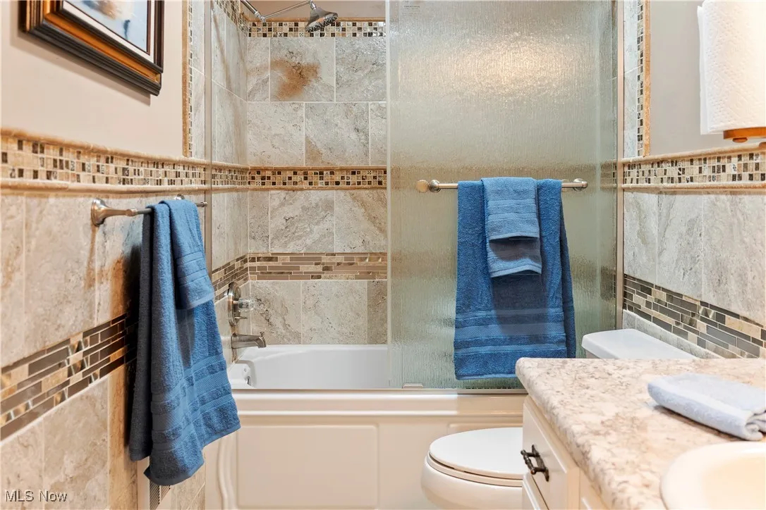 Bathroom featuring shower / bath combination with glass door, tile walls, toilet, and​​‌​​​​‌​​‌‌​‌‌​​​‌‌​‌​‌​‌​​​‌​​ vanity