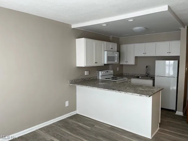Kitchen featuring a sink, white appliances, baseboards, a peninsula, and dark wood-style flooring