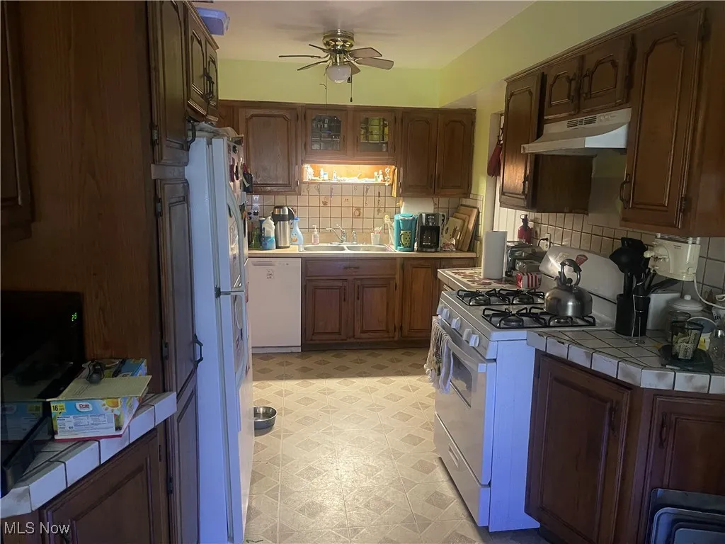 Kitchen featuring white appliances, under cabinet range hood, tile counters, ceiling fan, and decorative backsplash