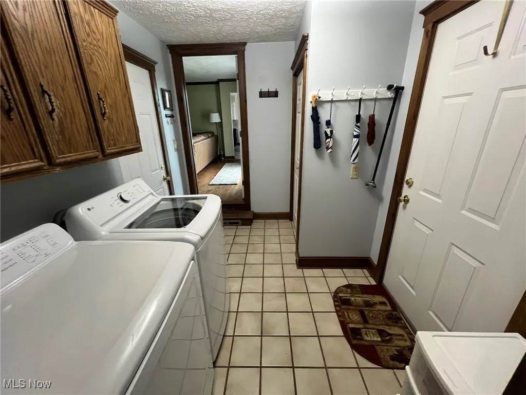 Clothes washing area with washing machine and clothes dryer, a textured ceiling, baseboards, cabinet space, and light tile patterned floors