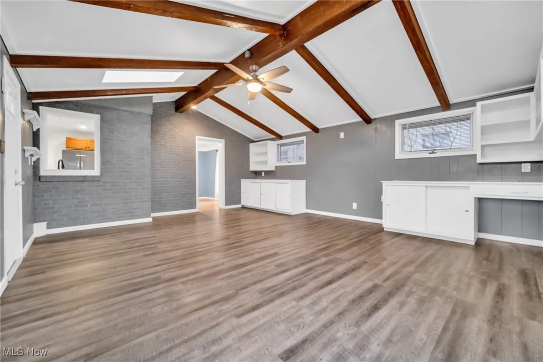 Unfurnished living room with baseboards, vaulted ceiling with skylight, brick wall, a ceiling fan, and wood finished floors