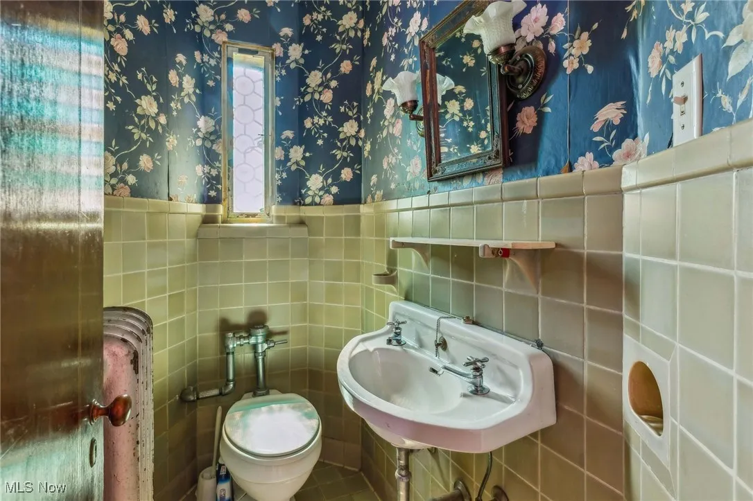 Half bathroom featuring tile walls, wallpapered walls, a wainscoted wall, and a sink