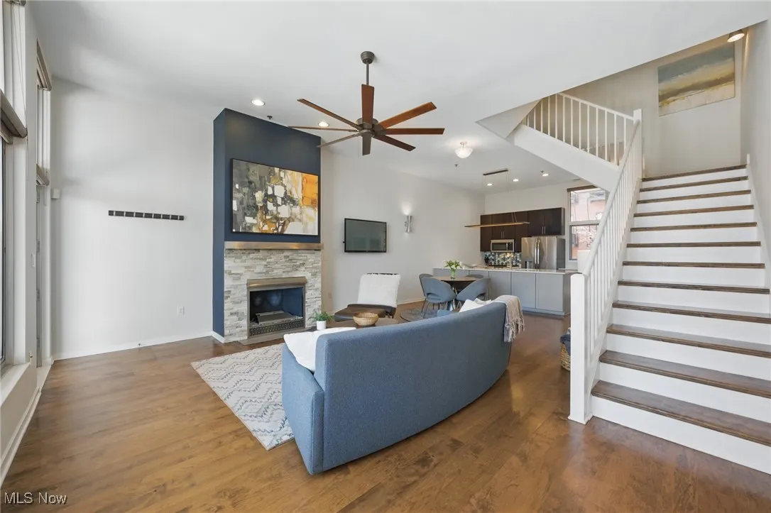 Living area with a stone fireplace, dark wood-style flooring, stairs, and ceiling fan