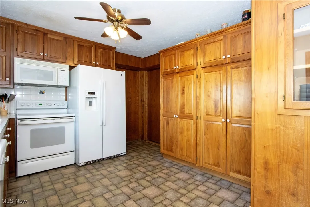 Kitchen featuring white appliances, brown cabinetry, light countertops, ceiling fan, and brick patterned floor