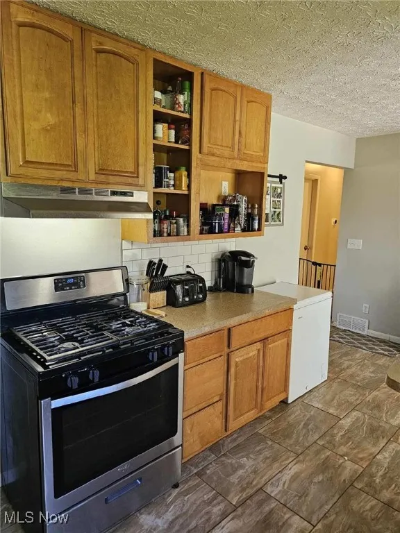 Kitchen featuring brown cabinetry, open shelves, stainless steel gas stove, under cabinet range hood, and tasteful backsplash