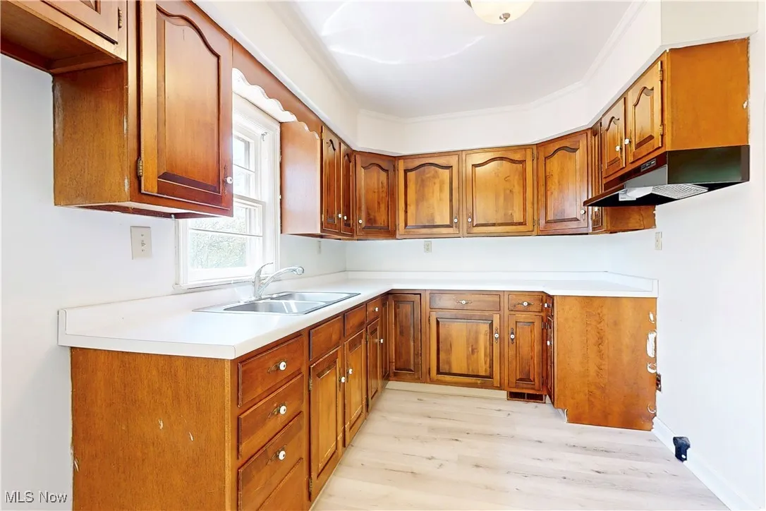 Kitchen featuring a sink, under cabinet range hood, brown cabinetry, light wood-style flooring, and light countertops