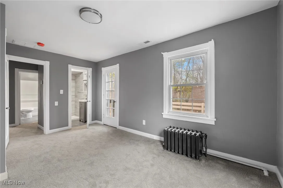 Unfurnished bedroom with connected bathroom, light carpet, and radiator heating unit