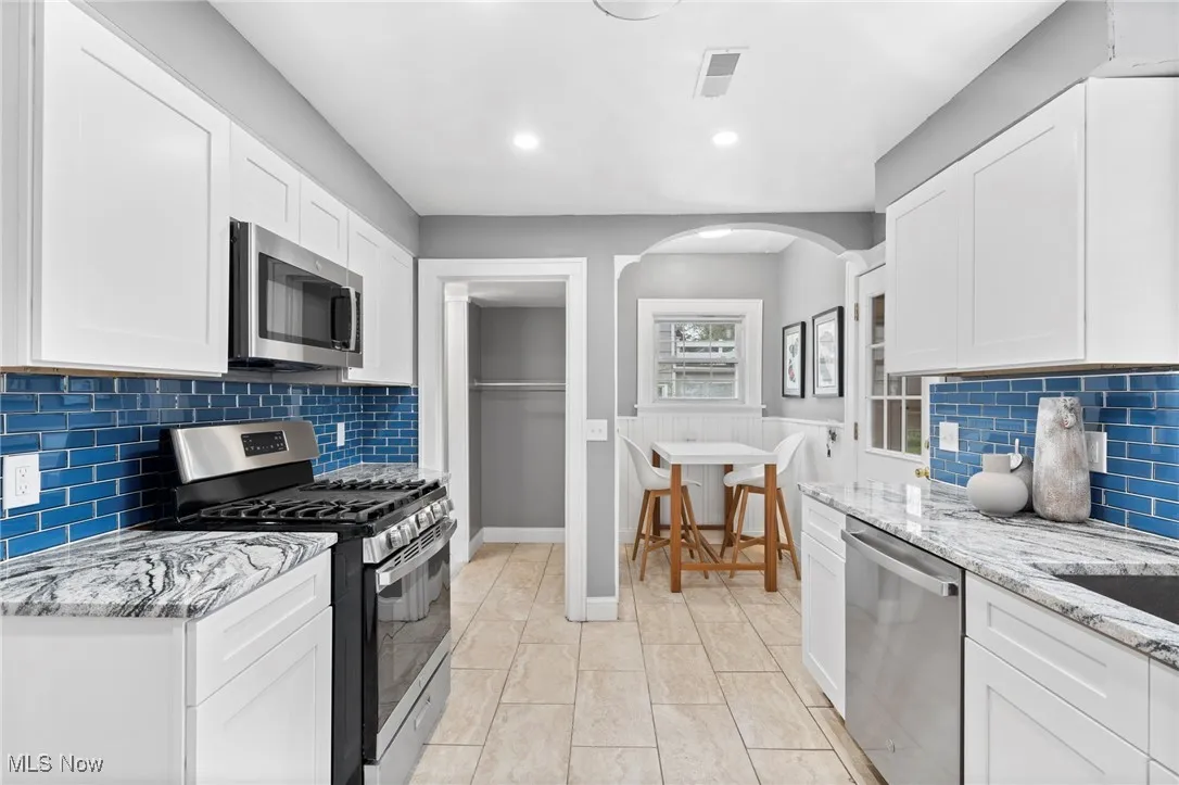 Kitchen featuring white cabinets, tasteful backsplash, light tile patterned floors, appliances with stainless steel finishes, and light stone counters