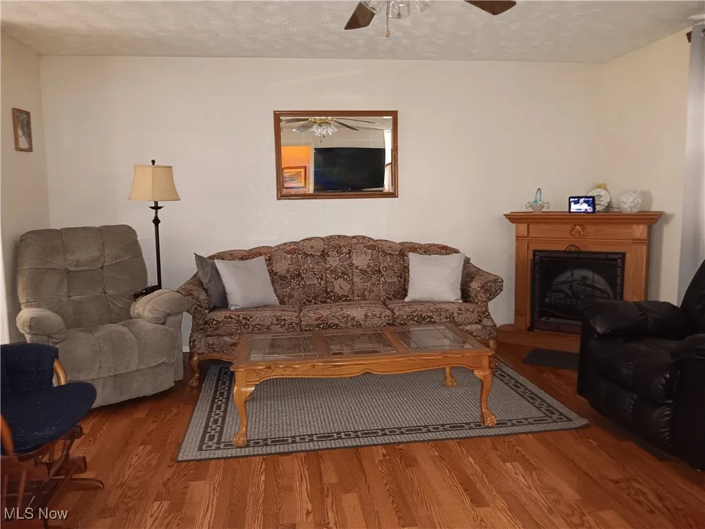 Living area with a textured ceiling, ceiling fan, a fireplace with raised hearth, and wood finished floors