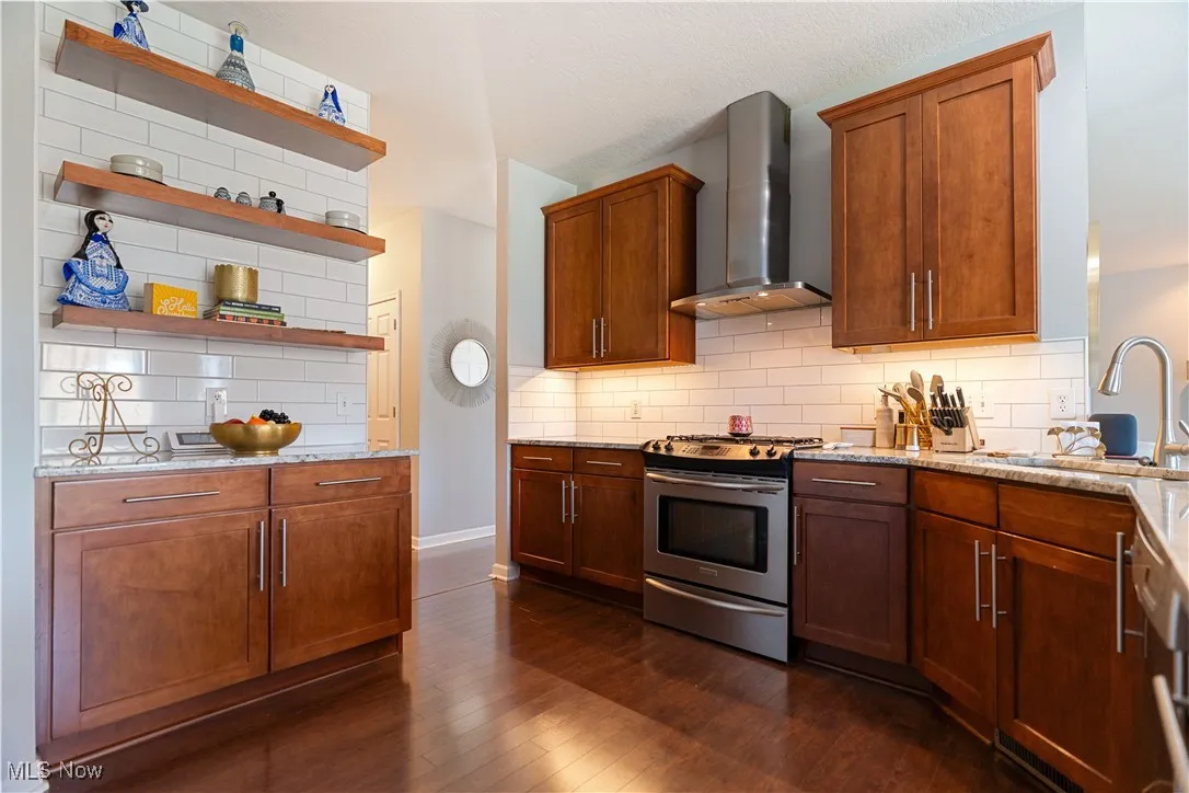 Kitchen featuring wall chimney range hood, open shelves, a sink, stainless steel gas range oven, and light stone countertops