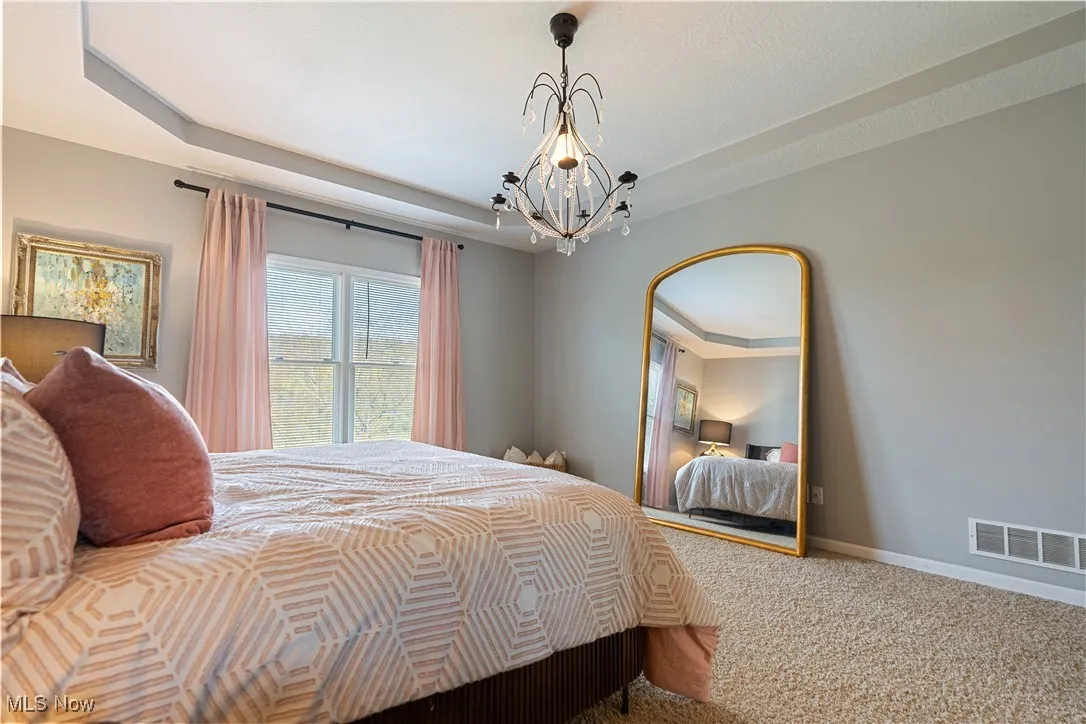 Carpeted bedroom featuring a notable chandelier, visible vents, a raised ceiling, and baseboards