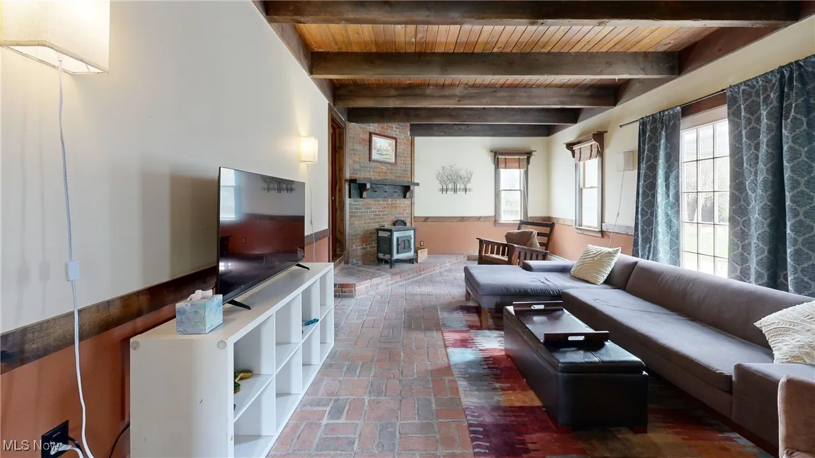 Living room featuring a wood stove, wooden ceiling, beam ceiling, brick floor, and wainscoting