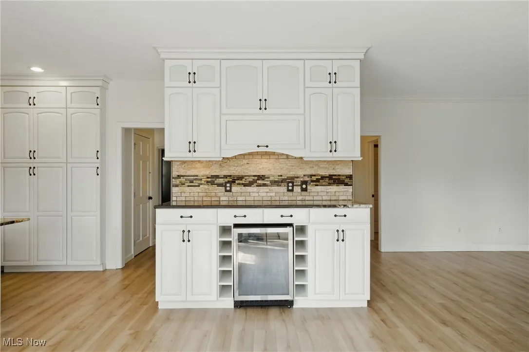 Kitchen with beverage cooler, dark countertops, light wood-style flooring, backsplash, and white cabinetry