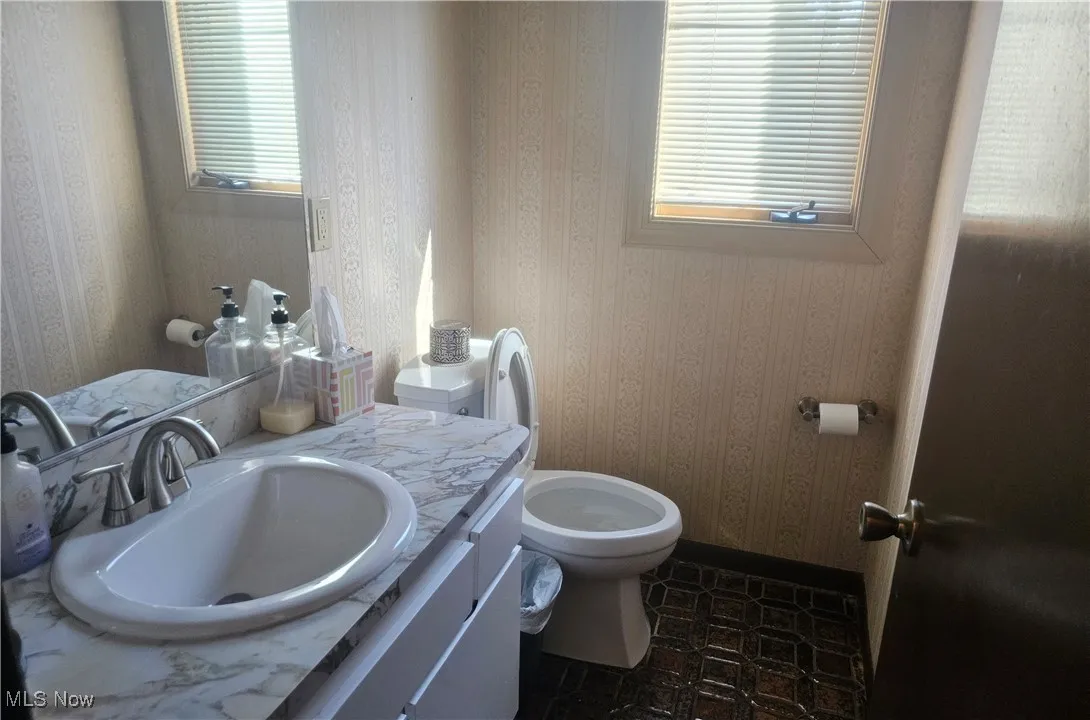 Half bathroom with toilet, wallpapered walls, baseboards, and vanity
