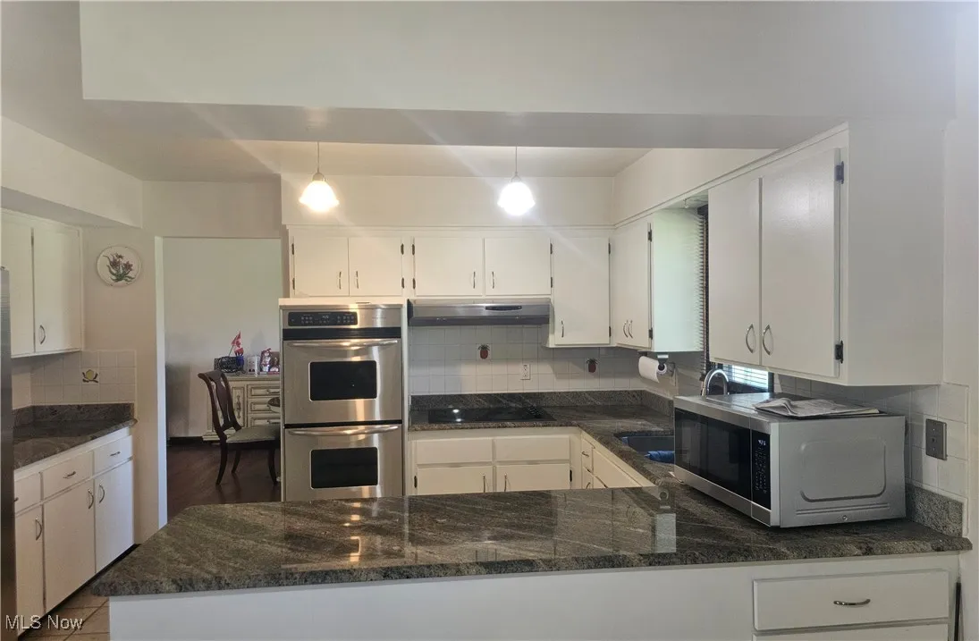 Kitchen featuring stainless steel double oven, dark stone counters, white cabinetry, under cabinet range hood, and tasteful backsplash