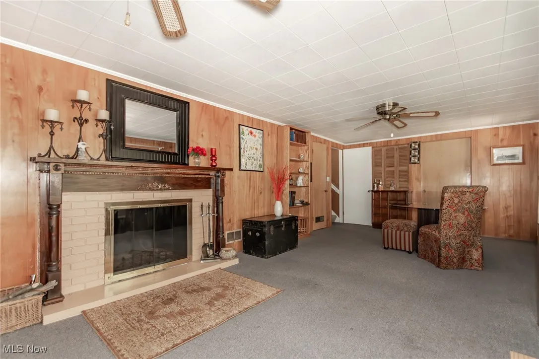 Carpeted living room featuring visible vents, a ceiling fan, a brick fireplace, and wooden walls