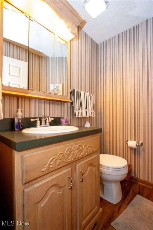 Half bath featuring vanity, toilet, a textured ceiling, wallpapered walls, and wood finished floors