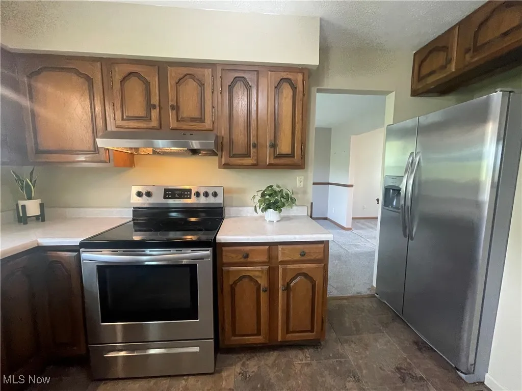 Kitchen with light countertops, appliances with stainless steel finishes, brown cabinetry, and under cabinet range hood