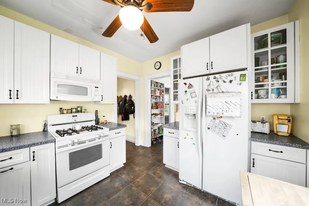 Kitchen with dark countertops, a ceiling fan, white cabinets, and white appliances